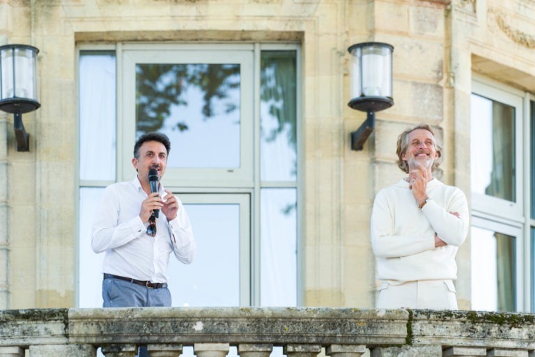 Deux hommes se tiennent sur un balcon en pierre avec une grande fenêtre en arrière-plan lors de la Soirée annuelle du Groupe Betclic. L'homme à gauche, parlant dans un micro, porte une chemise blanche avec un pantalon gris. L'homme à droite, vêtu d'une tenue entièrement blanche, sourit avec sa main sur son menton. Deux appliques murales sont visibles derrière eux.