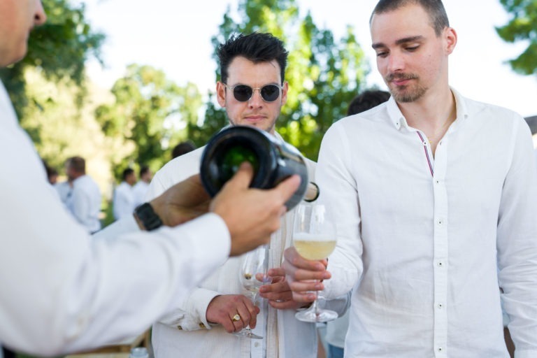 Deux hommes en chemise blanche tiennent des verres à vin lors de la Soirée annuelle du Groupe Betclic, un rassemblement en plein air. Une troisième personne, partiellement visible, verse du champagne d'une bouteille dans le verre d'un homme. Le décor est composé d'une végétation luxuriante et de personnages flous en arrière-plan, ce qui indique un événement social dans un cadre naturel.