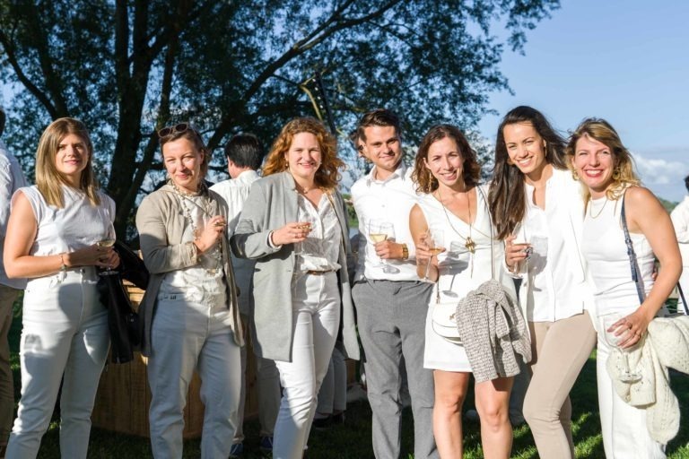 Un groupe de sept personnes, quatre femmes et trois hommes, se tiennent dehors sur l'herbe, vêtus de vêtements clairs et semi-formels. Ils sourient et tiennent un verre, avec des arbres et un ciel dégagé en arrière-plan. L'ambiance est joyeuse et ensoleillée, laissant présager d'un événement festif ou festif à la Soirée annuelle du Groupe Betclic.