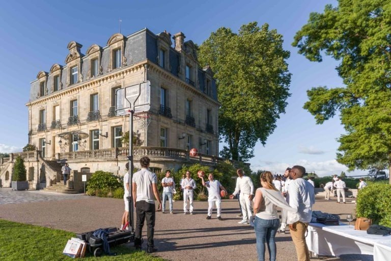 Un groupe de personnes vêtues de blanc jouent à un jeu ressemblant à du basket-ball devant un grand bâtiment décoré avec un toit en ardoise et des lucarnes. La scène, qui fait partie de la Soirée annuelle du groupe Betclic, se déroule par une journée ensoleillée avec des arbres à proximité. Quelques spectateurs et des sacs d'équipement éparpillés sont visibles autour de la scène.