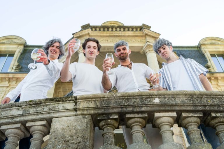 Quatre jeunes hommes se tiennent sur un balcon en pierre orné, tenant joyeusement des verres de vin blanc. Vêtus de chemises blanches décontractées, ils célèbrent la soirée annuelle du groupe Betclic dans le décor d'un élégant bâtiment historique avec de grandes fenêtres et un toit en pente. La photo est prise en contre-plongée, soulignant la grandeur du cadre.