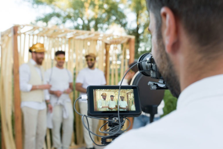 Un vidéaste filme trois hommes debout devant une structure en bois ornée de rubans dorés lors de la Soirée annuelle du Groupe Betclic. Les hommes sourient, sont vêtus de chemises blanches et portent des chapeaux dorés et des lunettes de soleil. Le viseur montre le trio en gros plan, sur un fond de feuillage vert.