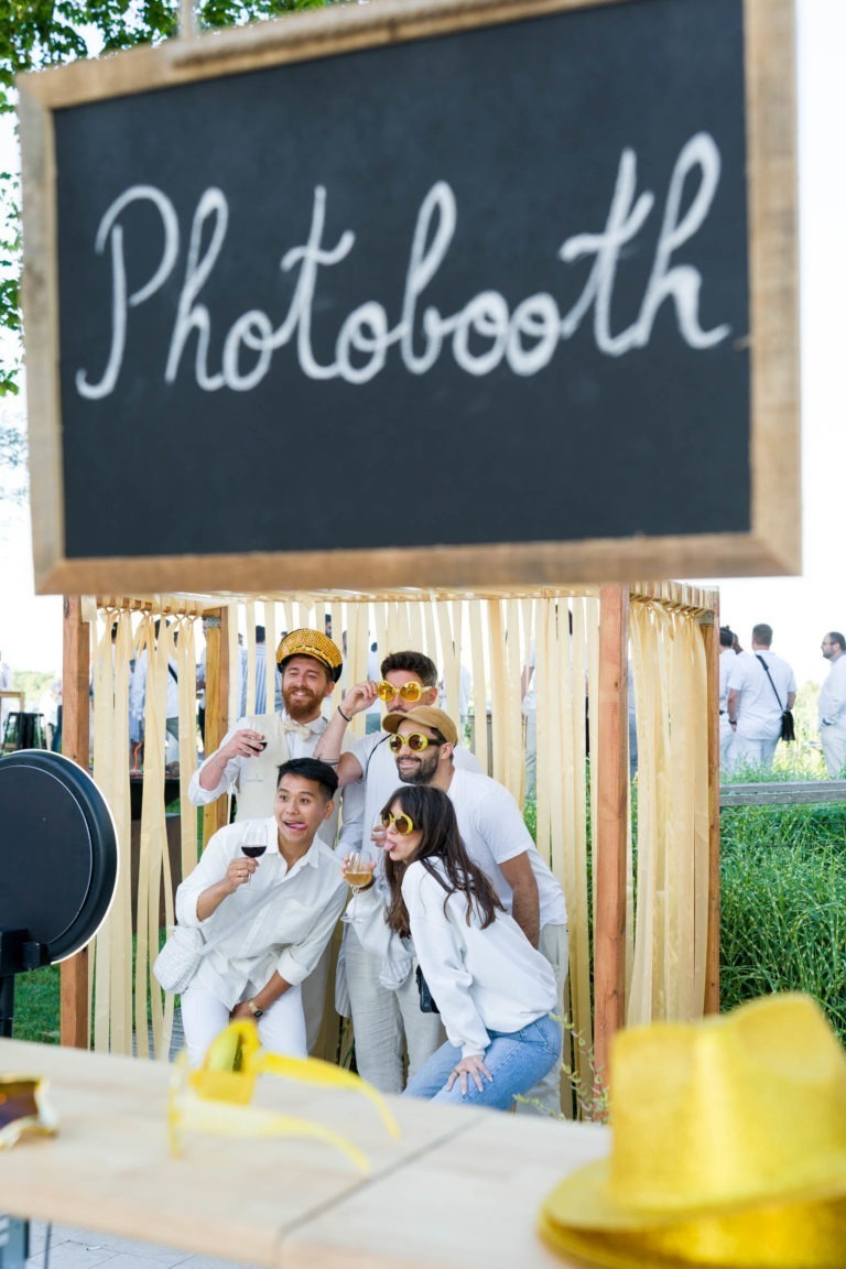 Un groupe de cinq personnes pose pour une photo dans un photomaton lors de la Soirée annuelle du Groupe Betclic. Elles portent toutes des tenues blanches et des accessoires amusants, notamment des chapeaux, des lunettes et des articles de fantaisie. Le photomaton est décoré de banderoles jaunes et un panneau en ardoise au-dessus indique « Photobooth ». L'ambiance est joyeuse.