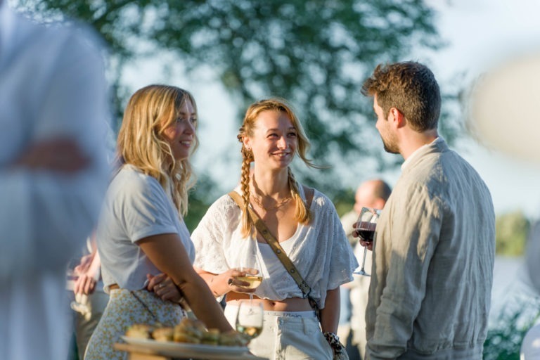 Trois personnes sont réunies à l'extérieur, en pleine conversation lors d'un événement social en plein jour. Deux femmes et un homme, vêtus de vêtements d'été légers, tiennent des boissons et sourient. Un arbre et d'autres invités flous sont visibles en arrière-plan, suggérant une atmosphère extérieure et détendue à la Soirée annuelle du Groupe Betclic.