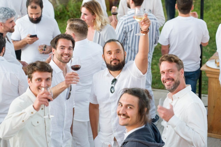 Un groupe d'hommes vêtus de blanc se rassemblent à l'extérieur, souriant et tenant des boissons lors de la soirée annuelle du groupe Betclic. Un homme au milieu lève un verre de vin blanc. L'arrière-plan montre d'autres personnes vêtues de blanc, certaines en train de converser, l'une utilisant un smartphone. Le décor respire clairement une atmosphère de fête.