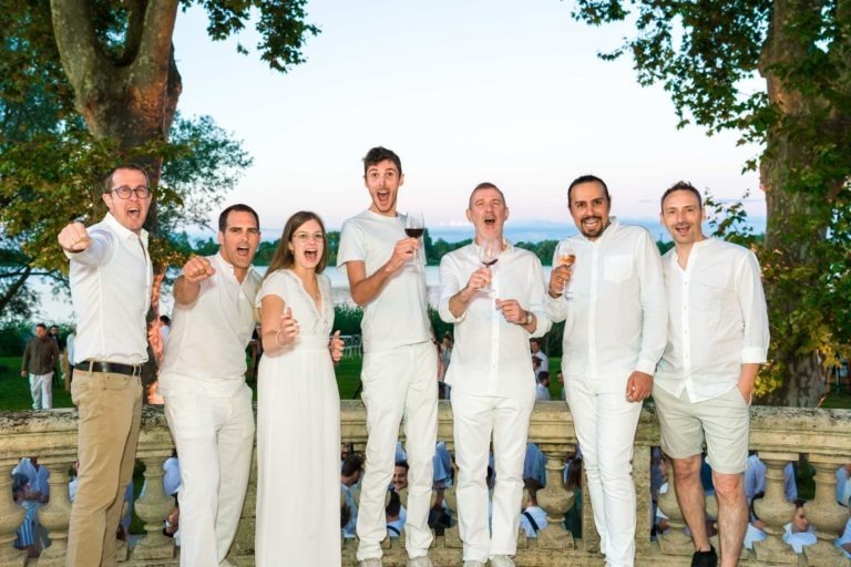 Un groupe de sept personnes, quatre hommes et trois femmes, se tiennent à l'extérieur devant une balustrade en pierre, tous vêtus de blanc. Ils sourient, rient et certains tiennent des boissons. L'arrière-plan présente une végétation luxuriante et une vue sur un plan d'eau au coucher du soleil lors de la Soirée annuelle du Groupe Betclic.