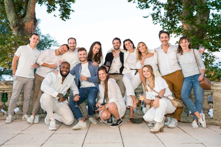 Un groupe de 14 personnes se tient debout ou accroupie à l'extérieur, tous souriant à la caméra. Ils sont habillés de façon décontractée, certains tenant un verre. Le décor semble être un parc avec des arbres et une balustrade en pierre derrière eux. Le ciel est clair, ce qui suggère un temps agréable pour la Soirée annuelle du Groupe Betclic.