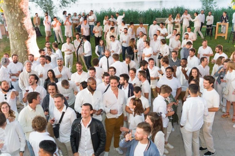 Un grand groupe de personnes vêtues de blanc ou de couleurs claires est rassemblé à l'extérieur près d'un plan d'eau. Certains tiennent des boissons et discutent tandis que d'autres sourient ou regardent la caméra. Le décor semble festif, représentant la Soirée annuelle du groupe Betclic, avec un mélange d'individus debout et assis profitant de l'événement.