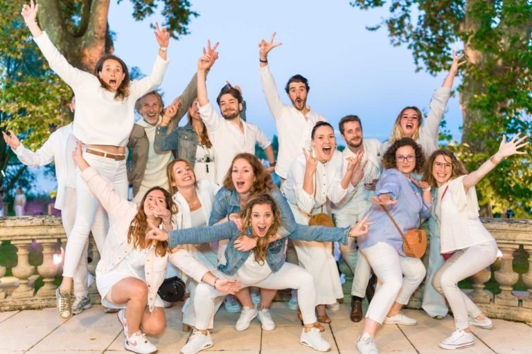 Un groupe joyeux de 17 personnes, la plupart vêtues de blanc, posent avec énergie dans un décor extérieur avec des arbres en arrière-plan. Ils affichent des expressions et des gestes ludiques, certains sautant ou s'accroupissant, créant une atmosphère animée et amusante. La toile de fond comprend une balustrade en pierre et un ciel clair, capturés lors de la Soirée annuelle du Groupe Betclic.