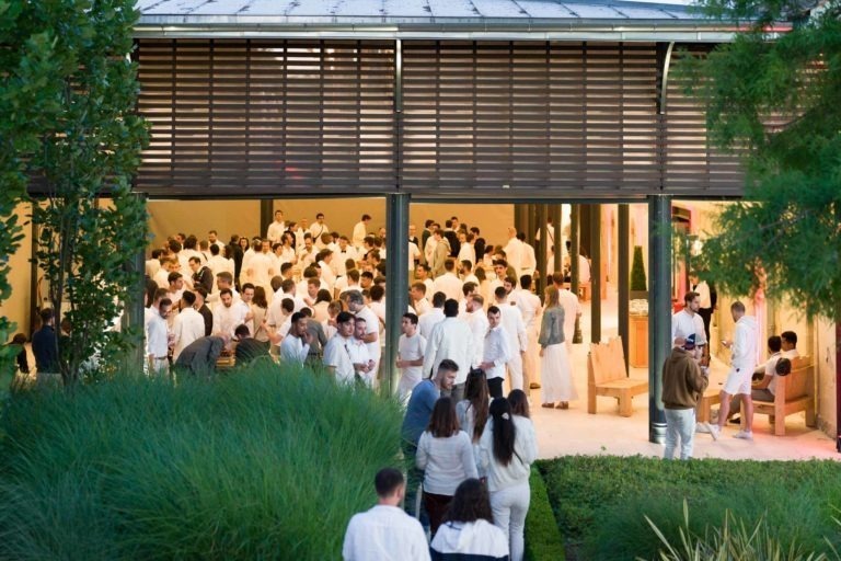 Un grand rassemblement de personnes vêtues de blanc est visible à travers les côtés ouverts d'un bâtiment aux lattes de bois verticales. L'espace est entouré d'une végétation luxuriante, avec quelques invités qui se mêlent à l'extérieur. L'espace intérieur semble lumineux, rempli de participants en train de socialiser à la Soirée annuelle du Groupe Betclic.