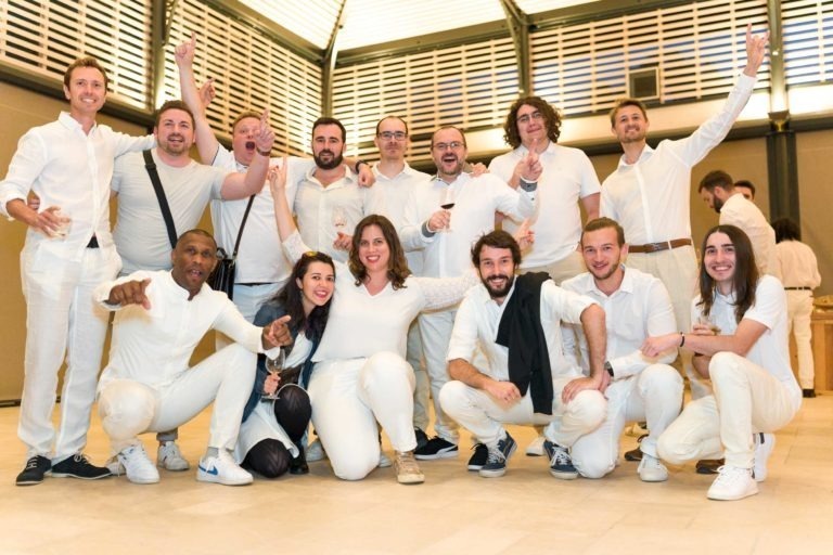 Un groupe de quatorze personnes, vêtues de blanc, posent joyeusement pour une photo lors de la Soirée annuelle du Groupe Betclic. Elles sourient, applaudissent et font divers gestes de célébration. Certaines sont debout, d'autres à genoux devant, le tout sur fond de murs et de fenêtres blancs et beiges.