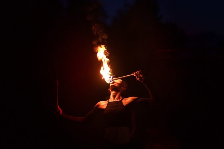 Lors de la Soirée annuelle du Groupe Betclic, un artiste dans un décor sombre tient une torche enflammée devant sa bouche ouverte, semblant cracher du feu. La flamme vive illumine son visage et le haut de son corps alors qu'il étend son autre bras vers l'extérieur avec une deuxième torche, le fond sombre ajoutant un effet dramatique.