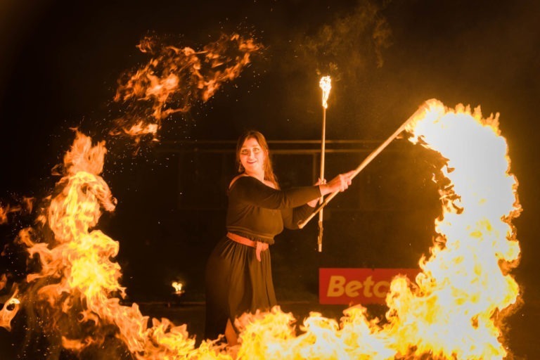 Une artiste vêtue de noir, tenant un bâton de feu avec des flammes, exécute une routine de danse du feu la nuit. Elle est entourée de flammes tourbillonnantes, créant un spectacle de lumière fascinant. Le fond est sombre, soulignant l'éclat des flammes. Un panneau avec les mots « Soirée annuelle Betclic Group » est partiellement visible.