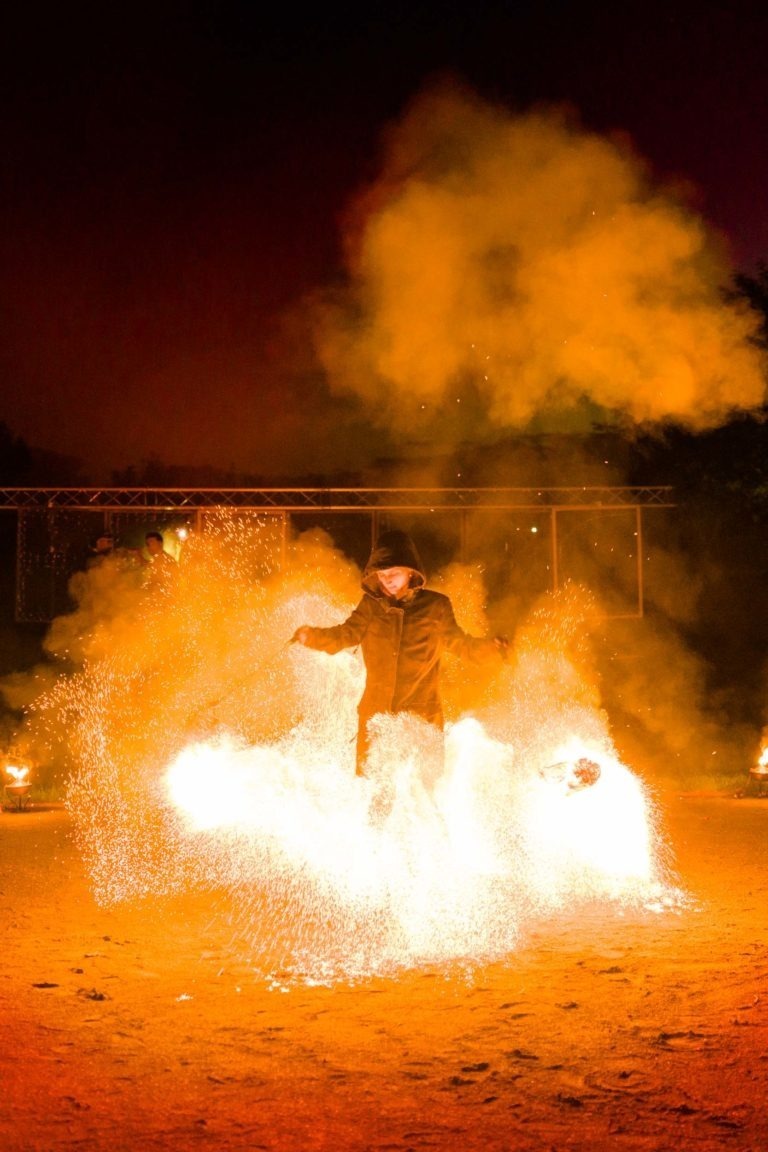 Une personne portant un manteau à capuchon exécute un numéro de feu lors de la Soirée annuelle du Groupe Betclic, entourée d'éclats de flammes et d'étincelles vibrantes. Des flammes orange et jaunes et de la fumée s'élèvent de manière spectaculaire dans le ciel sombre. Le sol semble avoir dispersé des étincelles, ajoutant à l'intensité de la performance enflammée.