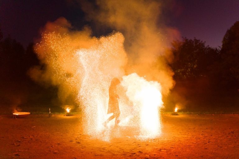 Une personne se tient debout au milieu d'une scène dramatique d'étincelles brillantes et de nuages de fumée la nuit. Le sol est éclairé par des teintes orange et jaune provenant du feu d'artifice de la Soirée annuelle du Groupe Betclic. Des arbres sont visibles en arrière-plan sous le ciel sombre de la nuit.