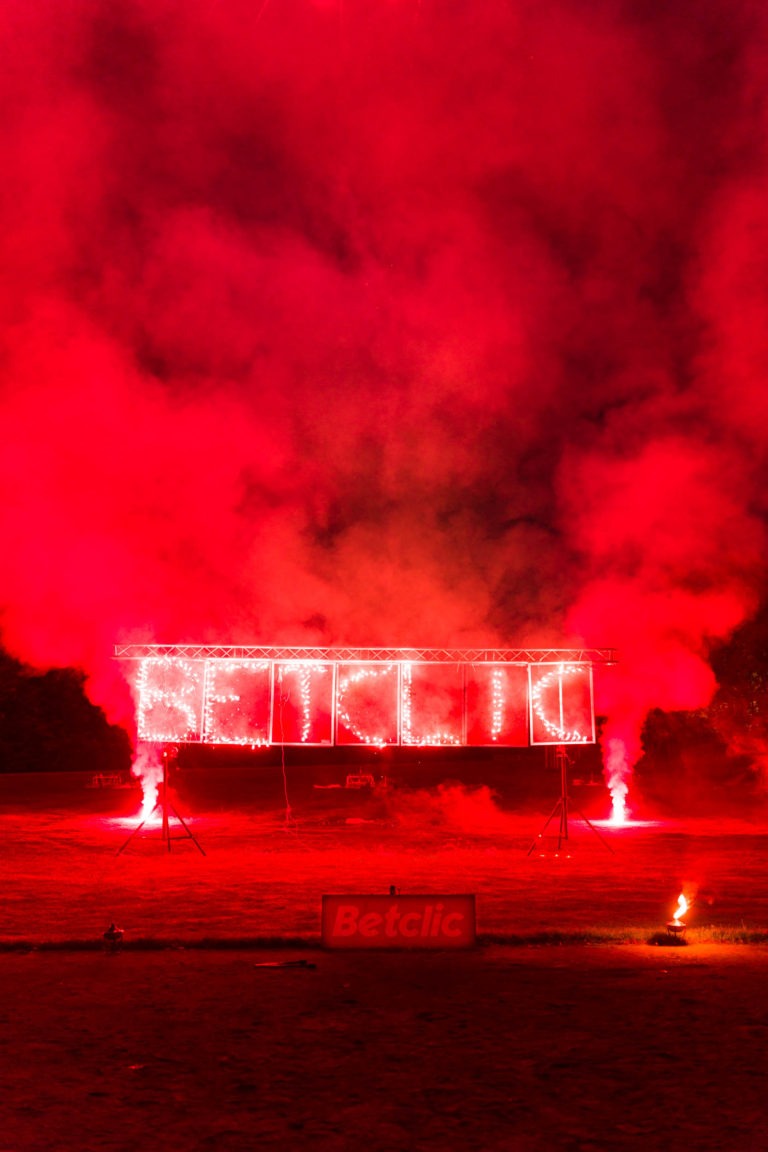 Une scène nocturne avec un spectacle pyrotechnique avec des fusées rouges et de la fumée à la Soirée Annuelle du Groupe Betclic. Des lettres lumineuses d'un rouge ardent forment le mot "Betclic" sur un cadre métallique. En dessous, un panneau de marque "Betclic" est visible. L'arrière-plan apparaît sombre, avec quelques arbres à peine visibles. L'ambiance générale est dramatique et intense.