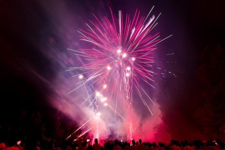 Un feu d'artifice exubérant illumine le ciel nocturne avec des éclats de lumière rouge, rose et blanche vibrants lors de la soirée annuelle du Groupe Betclic. Une grande foule de spectateurs se profile au premier plan, capturant la scène impressionnante tandis que des traînées de fumée persistent dans l'air, ajoutant à l'atmosphère dramatique.