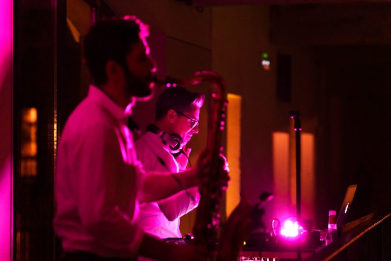 Lors de la soirée annuelle du Groupe Betclic, un DJ et un saxophoniste se produisent dans un décor illuminé aux lumières roses et violettes. Le DJ, portant un casque, manipule le matériel, tandis que le saxophoniste, qui est au premier plan et légèrement flou, joue de son instrument. Un ordinateur portable et d'autres équipements de DJ sont visibles sur une table.