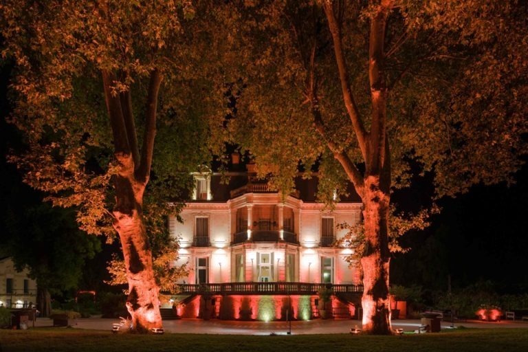 Derrière deux grands arbres se dresse un grand manoir illuminé de rouge et de blanc, qui accueille la soirée annuelle du groupe Betclic. Le bâtiment est doté de plusieurs balcons, de hautes fenêtres et de détails architecturaux décoratifs. L'éclairage spectaculaire crée une atmosphère enchanteresse, mettant en valeur à la fois le manoir et les arbres environnants.