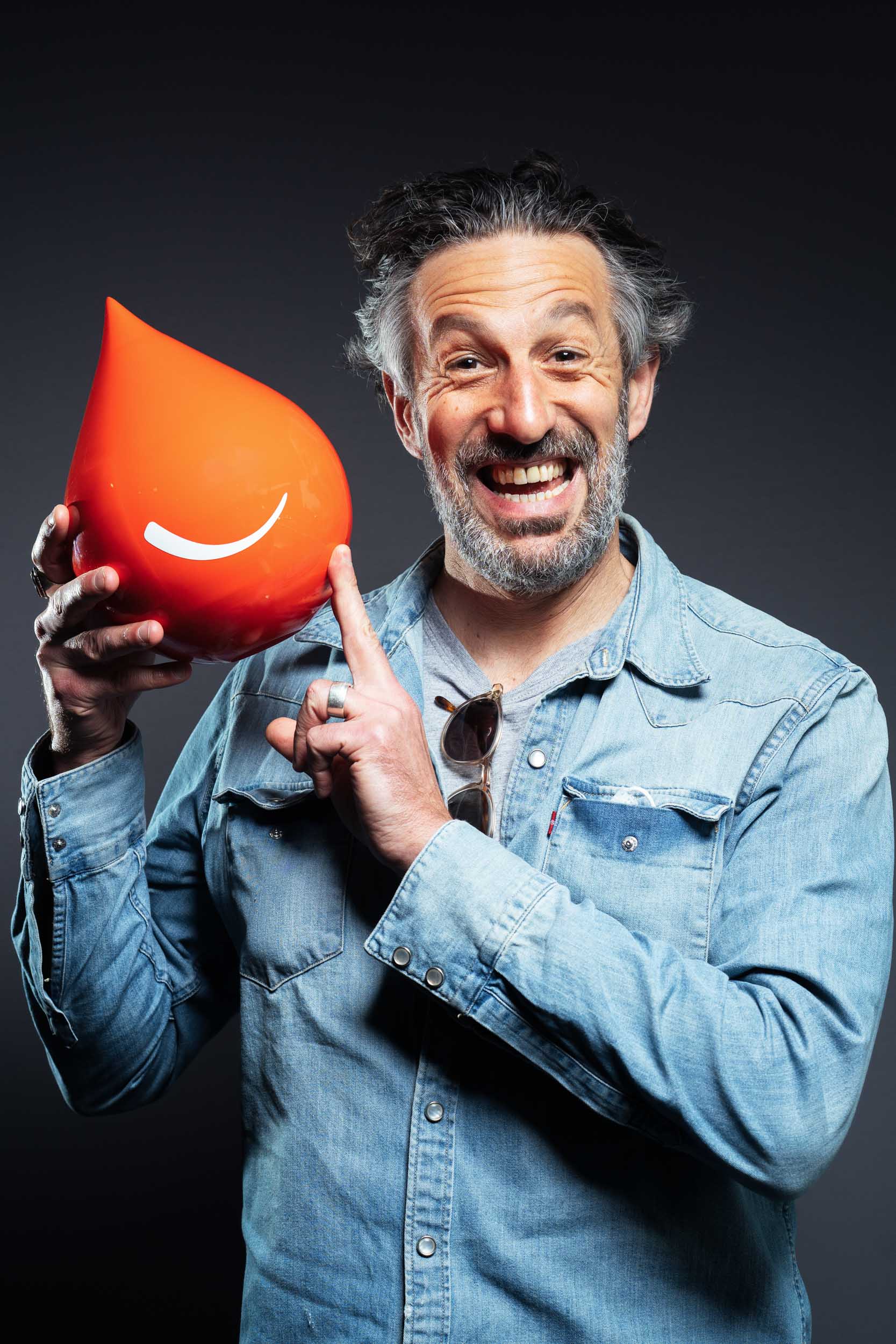 Un homme joyeux aux cheveux et à la barbe gris, vêtu d'une chemise en jean bleu clair, tient un grand objet rouge en forme de larme avec un smiley blanc dessus. L'homme sourit largement et pointe l'objet avec son autre main. L'arrière-plan est un dégradé sombre, parfait pour les portraits professionnels à Bordeaux.