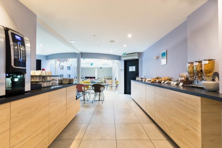 Un espace petit-déjeuner buffet bien éclairé dans une salle à manger avec des comptoirs en bois et noirs aux Appartements De Vacances Bordeaux. On y trouve des distributeurs de céréales, une machine à café sur la gauche et des produits de boulangerie exposés. Le coin salon comprend des chaises et des tables colorées, avec un espace lumineux et une télévision murale en arrière-plan.