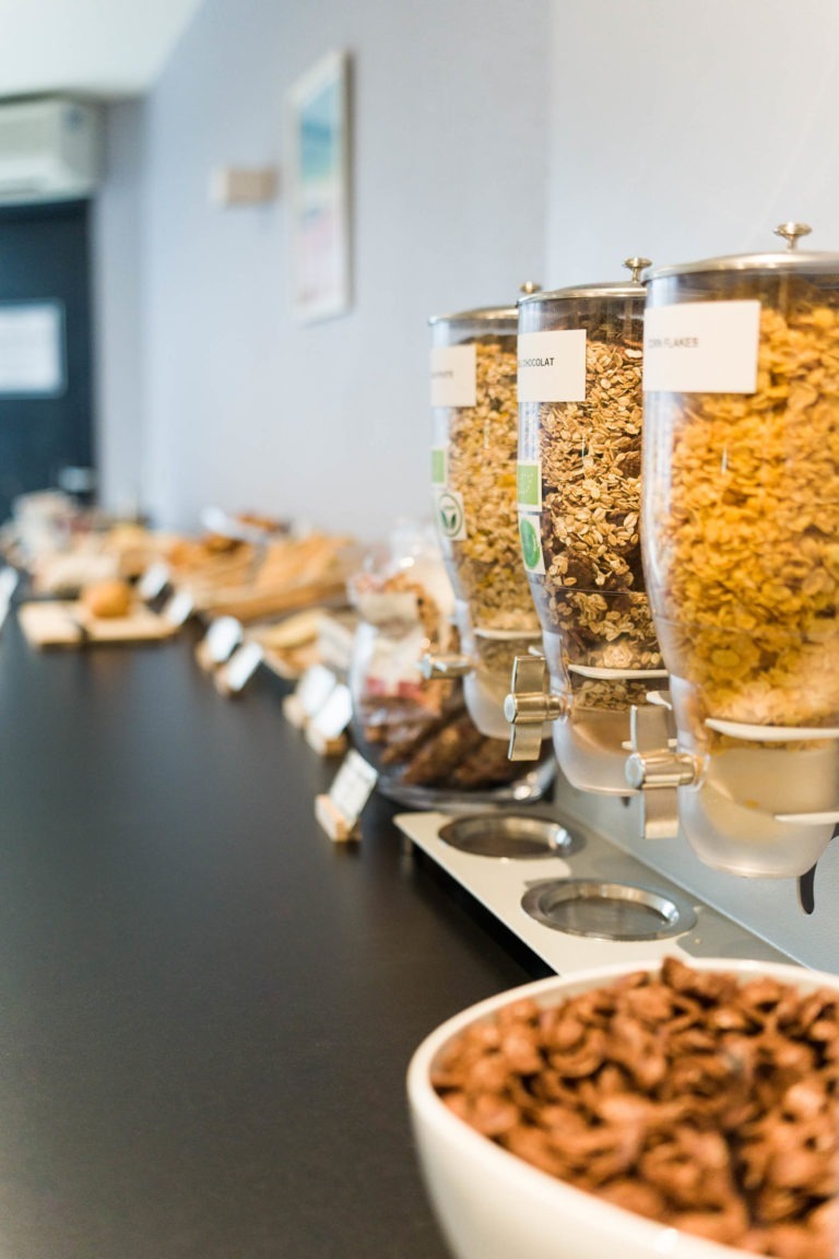 Une table de petit-déjeuner buffet aux Appartements De Vacances Bordeaux comporte plusieurs récipients proposant des céréales comme du granola et des cornflakes. Au premier plan, il y a un bol blanc rempli de céréales au chocolat. L'arrière-plan montre un assortiment de pains, de pâtisseries et d'autres produits de petit-déjeuner exposés.