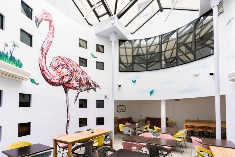 Un café lumineux et moderne avec de hauts plafonds et une grande lucarne. Les murs blancs sont ornés d'une superbe fresque murale représentant un flamant rose, évoquant le charme des Appartements De Vacances Bordeaux. Des chaises colorées et des tables en bois remplissent le coin salon, tandis que de petites fenêtres et des climatiseurs sont judicieusement placés sur les murs.