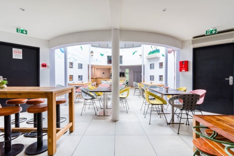 La salle à manger lumineuse et moderne des Appartements De Vacances Bordeaux est dotée de chaises colorées autour de tables en bois. À gauche se trouve une table haute avec des tabourets en bois et une colonne centrale ajoute du caractère. Les portes de sortie sont sur les côtés. De grandes fenêtres permettent à la lumière naturelle de remplir l'espace, créant ainsi une atmosphère ouverte.