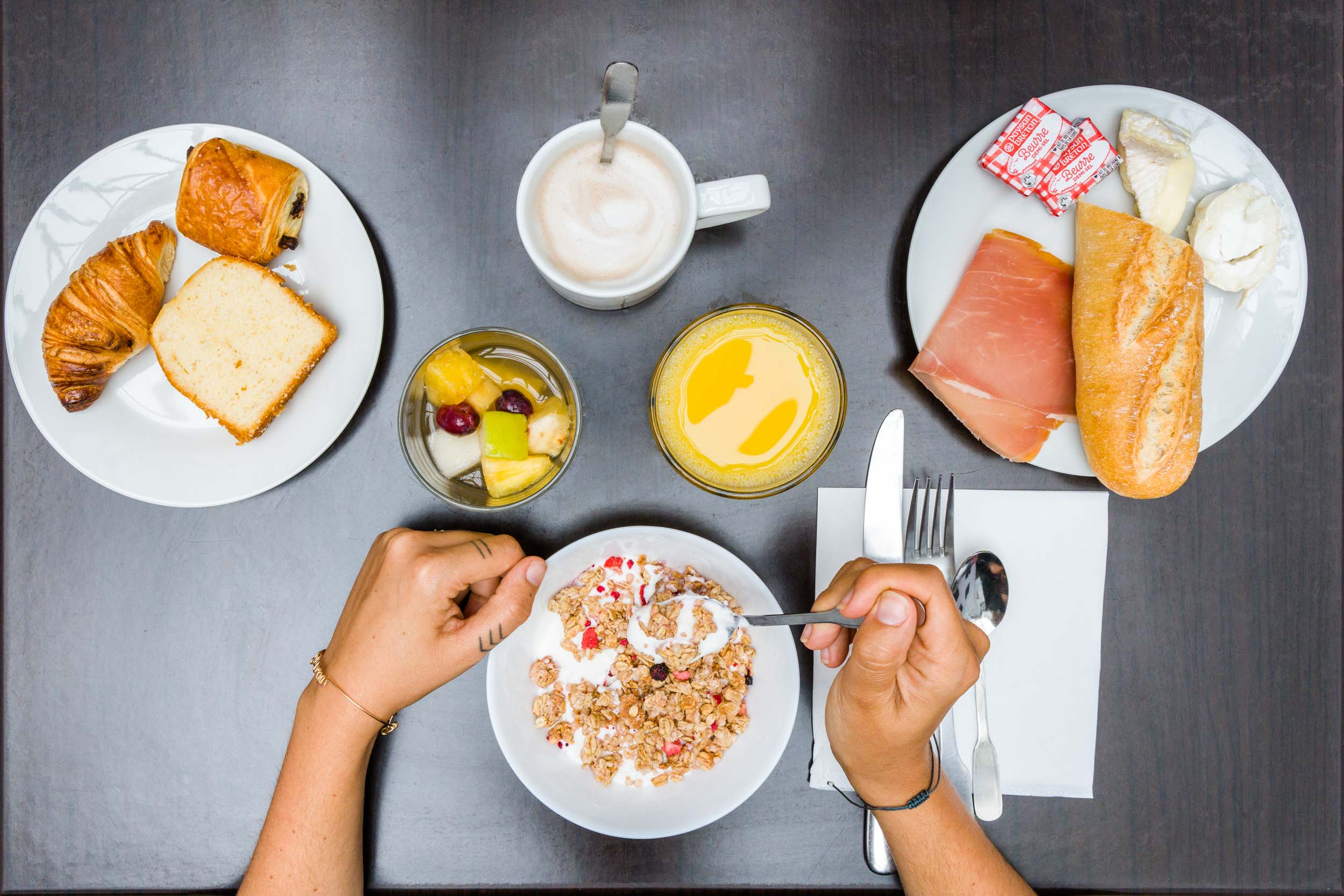 Une table de petit-déjeuner aux Appartements De Vacances Bordeaux propose différents plats : une personne déguste du granola avec du yaourt, des fruits frais et une boisson chaude, probablement du café ou du chocolat chaud. Autour du bol se trouvent des assiettes avec des pâtisseries, des tranches de pain, du jambon, de la mozzarella, du jus d'orange et un verre rempli de salade de fruits.