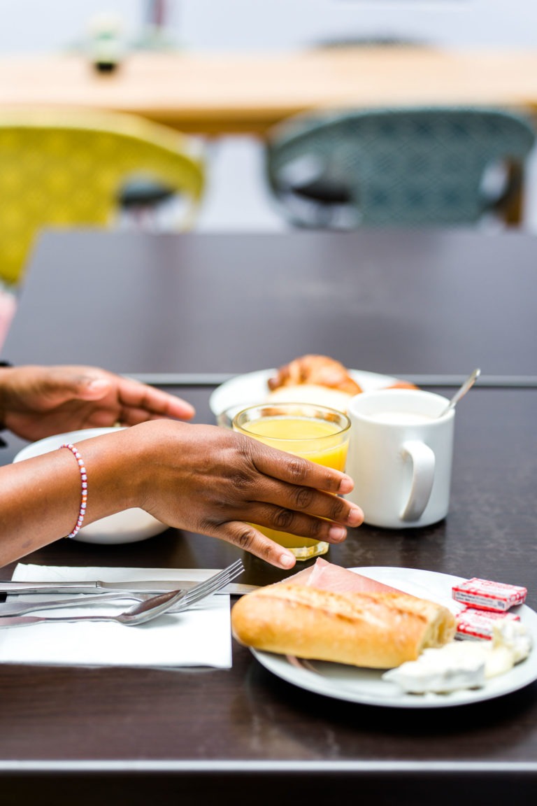 Une personne tient un verre de jus d'orange à une table avec une variété de produits pour le petit-déjeuner, rappelant les matinées passées aux Appartements De Vacances Bordeaux. Devant elle se trouve une assiette avec un croissant et des petits paquets de confiture, à proximité d'une tasse blanche, des ustensiles sur une serviette et une assiette avec une baguette et du fromage à pâte molle. Deux chaises sont visibles à l'arrière-plan.