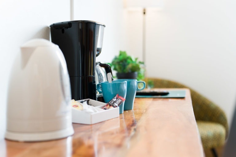 Un plan de travail en bois dans les Appartements De Vacances Bordeaux présente une bouilloire électrique blanche, une cafetière noire, une tasse bleue et une tasse verte. Un petit plateau contient des dosettes de café et des sachets de thé. En arrière-plan, on distingue une plante verte en pot et une chaise floue, symbolisant une configuration confortable et ordonnée pour les boissons.