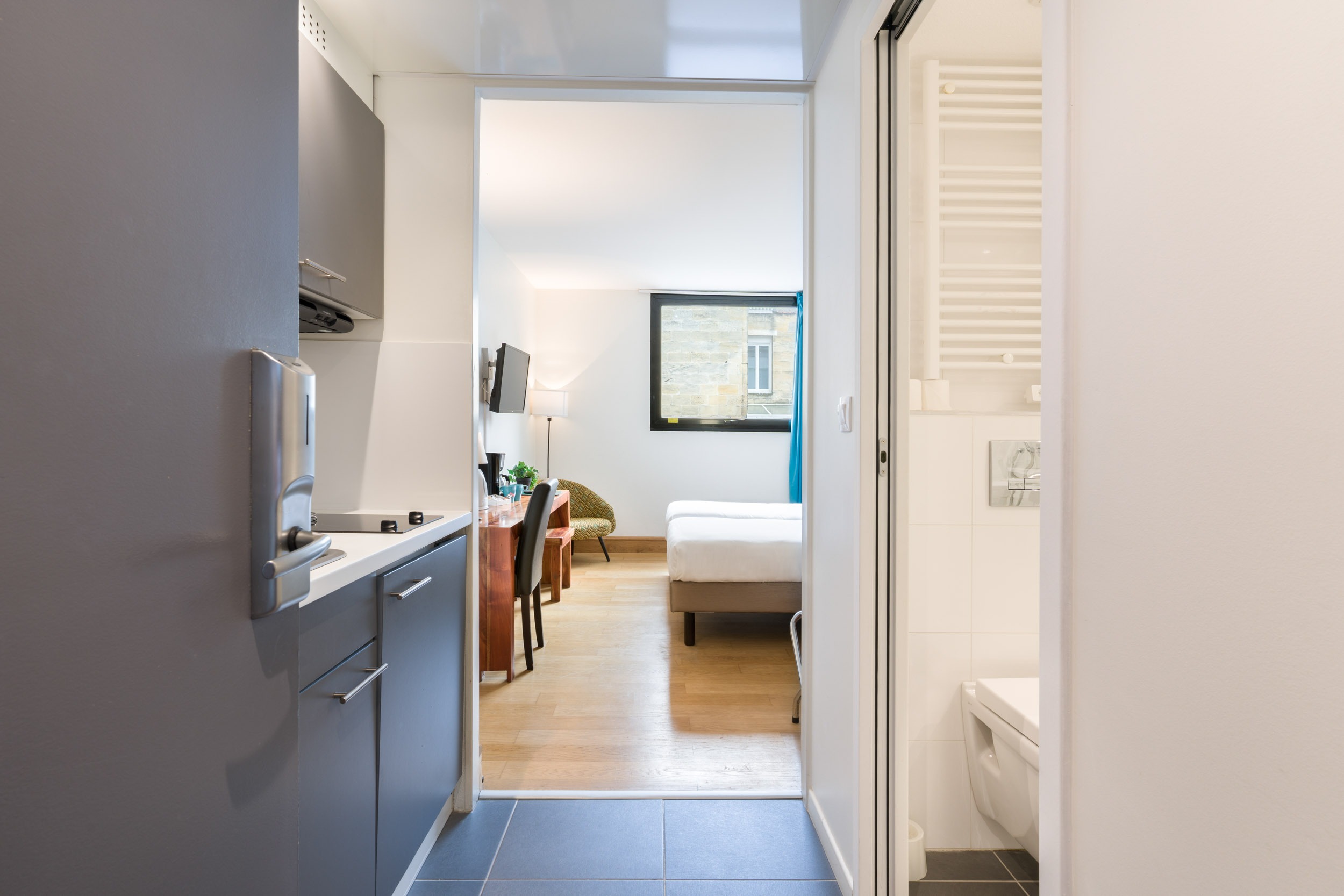 Un studio compact dans les charmants Appartements De Vacances Bordeaux offre une vue depuis l'entrée. Sur la gauche, il y a une petite kitchenette avec des placards, un évier et une cuisinière. Tout droit se trouve un coin salon avec un lit, un bureau, des chaises et une télévision murale. Sur la droite, une salle de bain carrelée de blanc est partiellement visible.