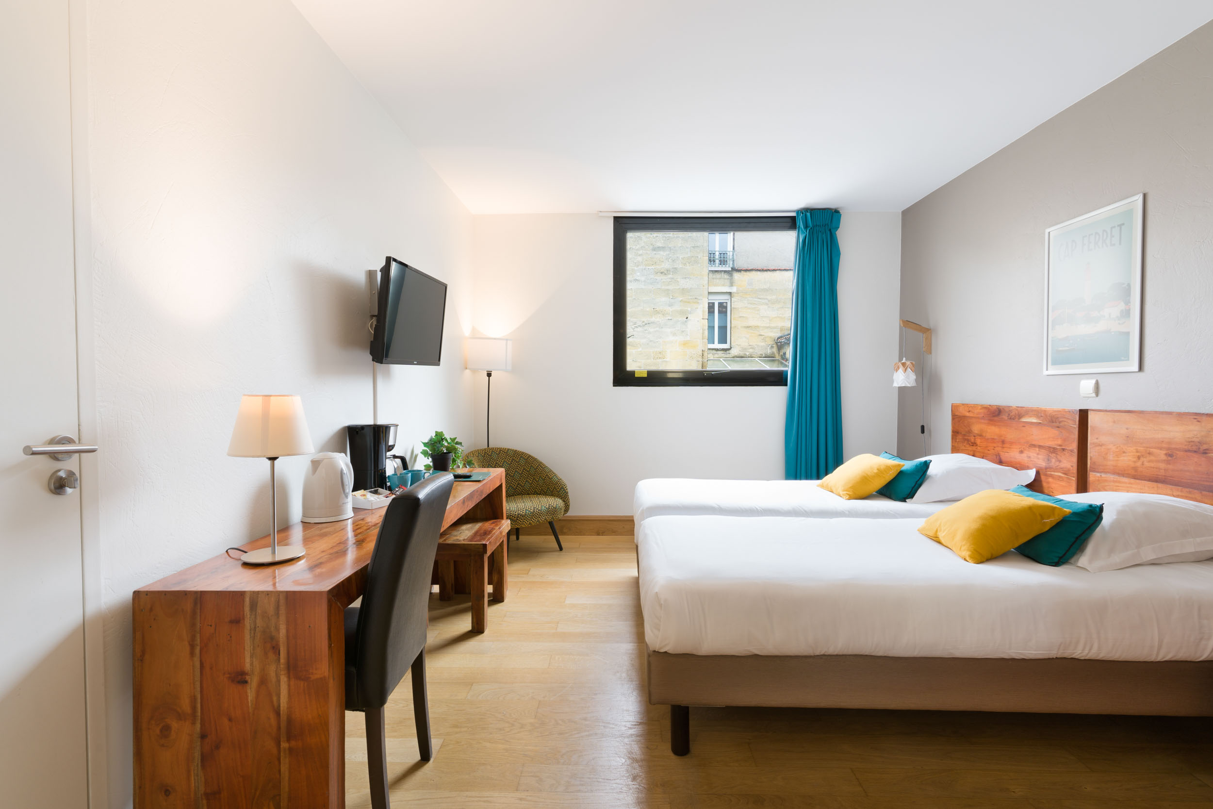 Une chambre d'hôtel moderne de l'Appartements De Vacances Bordeaux dispose de deux lits simples ornés de draps blancs et d'oreillers jaunes et bleus. À côté d'une télévision à écran plat fixée au mur, vous trouverez un bureau en bois avec une lampe, une chaise et un plateau/bouilloire. La chambre comprend une fenêtre avec des rideaux bleu canard, une chaise rembourrée verte et du parquet.