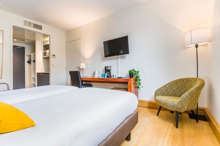 Une chambre d'hôtel moderne dans le style des Appartements De Vacances Bordeaux, dotée de deux lits jumeaux soigneusement confectionnés avec des draps blancs et un coussin jaune. La chambre comprend un bureau en bois avec une chaise, une télévision murale, une lampe sur pied, un fauteuil à motifs et une kitchenette en arrière-plan. La décoration est épurée et minimaliste avec des murs de couleur claire et du parquet.