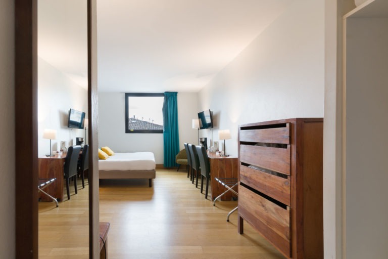 Cette chambre moderne de l'Appartements De Vacances Bordeaux est dotée de parquet et d'un lit double avec des draps blancs et deux oreillers jaunes. Deux chaises et un bureau avec lampes et miroirs se trouvent de chaque côté. La fenêtre est dotée de rideaux bleu sarcelle et une commode en bois se trouve sur le côté droit. Une télévision à écran plat est fixée au mur à côté du lit.