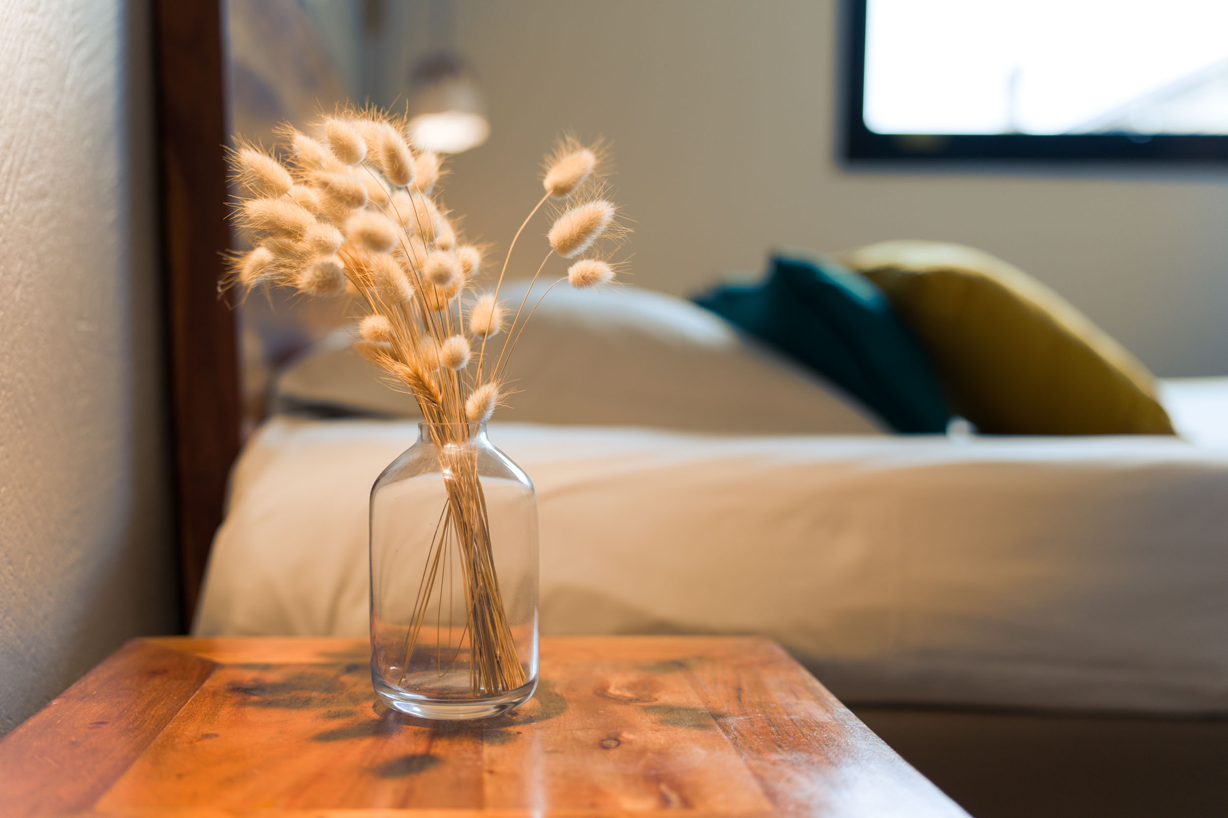 Un petit vase en verre rempli d'herbe à queue de lapin séchée est posé sur une table de chevet en bois dans l'un des charmants Appartements De Vacances Bordeaux. La table est à côté d'un lit avec une literie blanche et des oreillers colorés, avec une fenêtre en arrière-plan. La scène est doucement éclairée, créant une atmosphère chaleureuse et sereine.