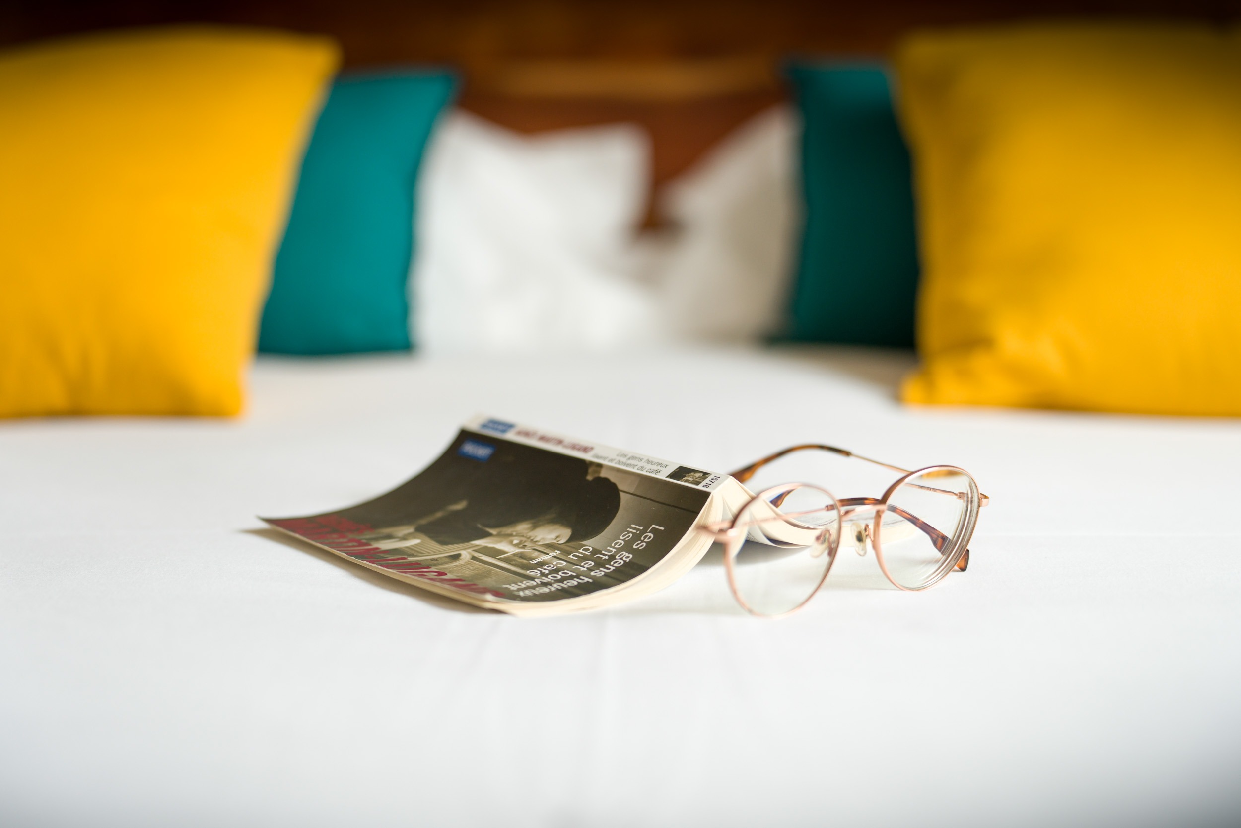 Une paire de lunettes repose sur un magazine posé sur un lit blanc dans l'un des confortables Appartements De Vacances Bordeaux. Le lit, orné de quatre oreillers vert canard et jaune moutarde, dégage une ambiance détendue et accueillante.