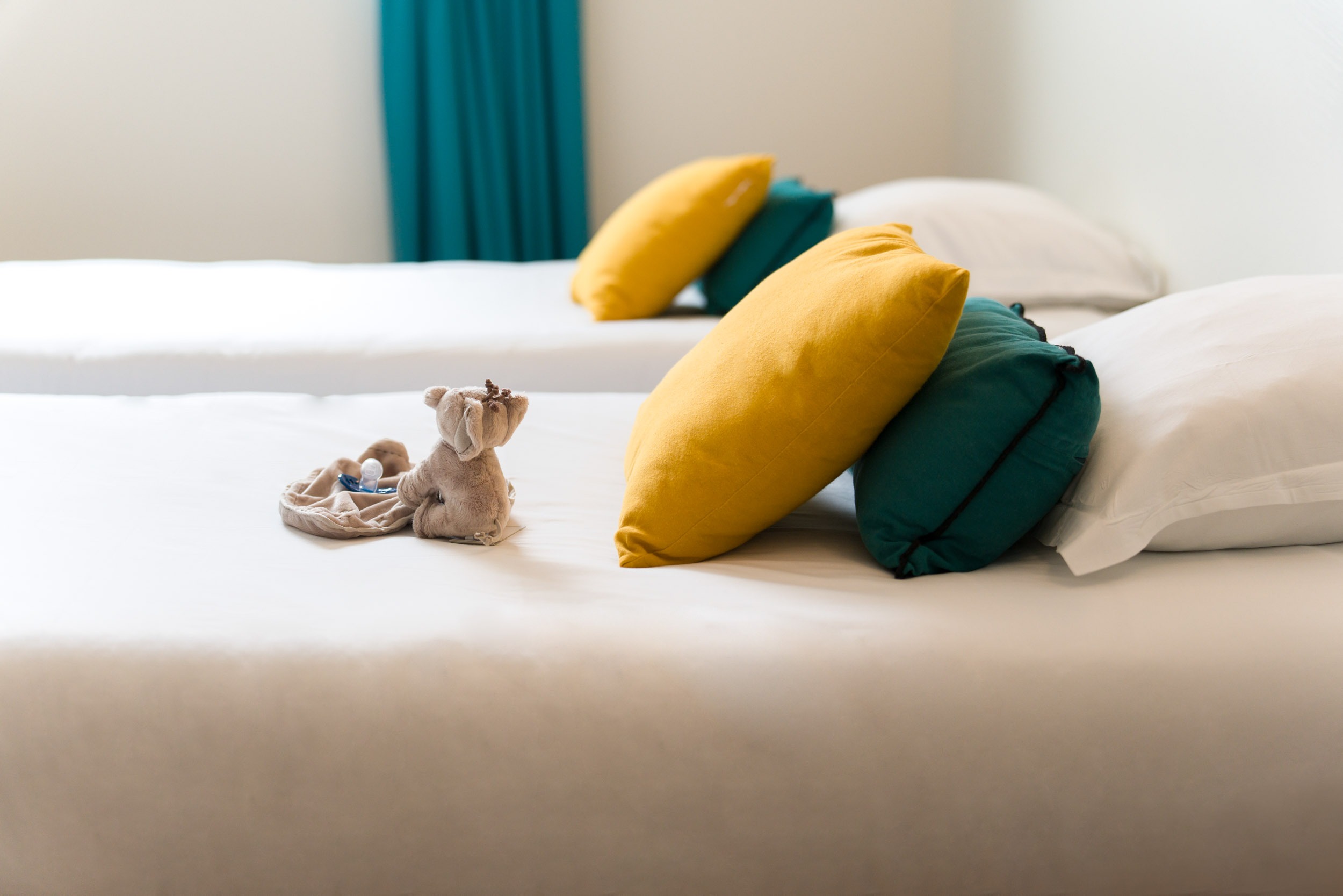 Un lit soigneusement fait avec des draps blancs est agrémenté de deux coussins colorés, l'un jaune et l'autre bleu sarcelle. Au pied du lit, un petit éléphant en peluche et une poupée reposent sur un fond en tissu beige. À l'arrière-plan, un deuxième lit fait écho au premier, créant ainsi une symétrie dans ces Appartements De Vacances Bordeaux.