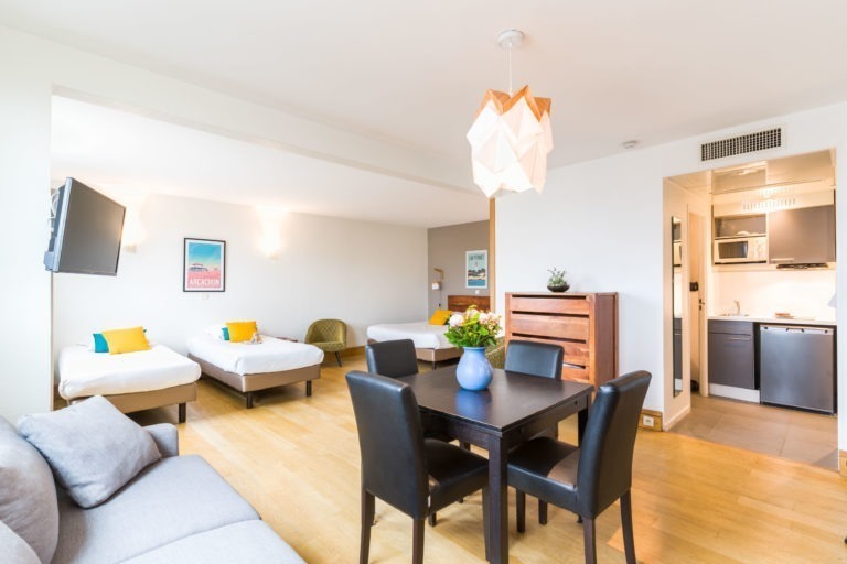 Un studio lumineux et moderne des Appartements De Vacances Bordeaux dispose de deux lits simples contre un mur blanc avec des coussins colorés, d'une petite télévision murale, d'un canapé, d'une table à manger noire avec quatre chaises, d'une commode et d'une petite cuisine équipée d'un micro-ondes et d'une cuisinière. Une suspension est suspendue au plafond.