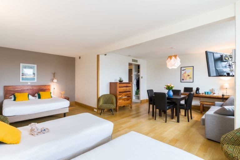 Une chambre spacieuse de l'hôtel Appartements De Vacances Bordeaux avec deux lits doubles, chacun orné de draps blancs et de coussins colorés. Une commode en bois et deux fauteuils sont installés près des lits. La chambre comprend un coin repas avec une table et des chaises noires, un canapé, une télévision murale et un espace de travail dans le coin.