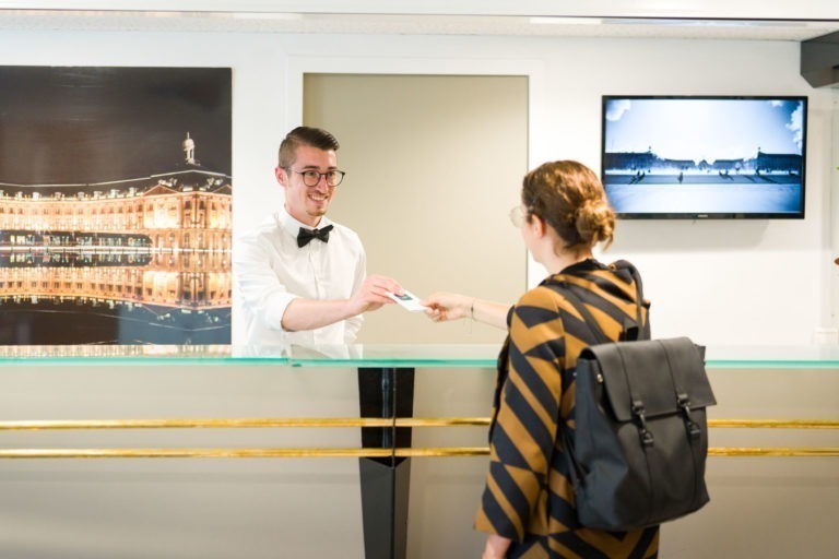 Une réceptionniste d'hôtel en chemise blanche et nœud papillon noir remet une carte-clé à un client portant un manteau rayé noir et jaune et un sac à dos noir. Derrière la réceptionniste, on voit une photo encadrée des Appartements De Vacances Bordeaux la nuit et un téléviseur fixé au mur affichant une image en noir et blanc.