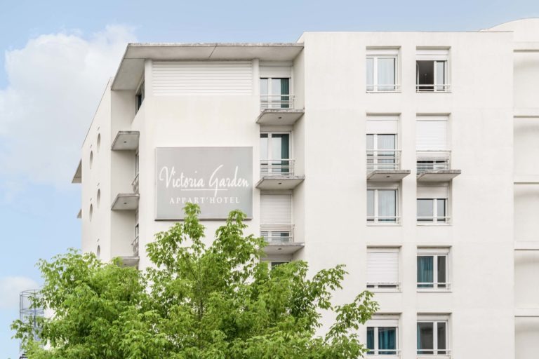 Un immeuble beige à plusieurs étages avec des balcons et de nombreuses fenêtres, certaines avec des volets fermés. Un panneau indiquant « Victoria Garden Appart Hotel » est bien visible sur le côté, mettant en valeur son attrait en tant qu'Appartements De Vacances Bordeaux. Des arbres verts sont au premier plan et le ciel est bleu avec de légers nuages.