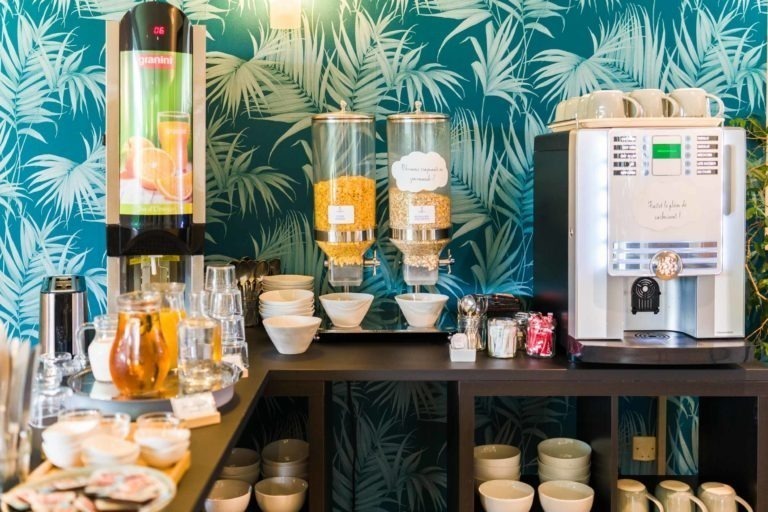 Le bar du petit-déjeuner des Appartements De Vacances Bordeaux est décoré d'un papier peint à thème tropical. Il y a deux distributeurs de céréales, un distributeur de jus d'orange et une machine à café. Différents bols, tasses, verres et assiettes sont soigneusement disposés. Une théière et une pile de condiments emballés sont également visibles sur le comptoir.