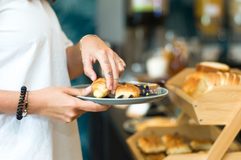 Une personne vêtue d'une chemise blanche tient une assiette bleu-gris et choisit une pâtisserie parmi un assortiment. Au premier plan, diverses pâtisseries sont disposées sur des étagères en bois. L'arrière-plan, légèrement flou, déplace l'attention vers les pâtisseries et les mains de la personne, créant une scène rappelant les matins dans les Appartements De Vacances Bordeaux.