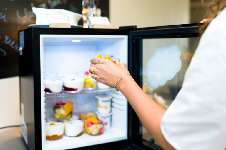 Une personne, les cheveux longs visibles, ouvre un petit réfrigérateur rempli de divers desserts dans les Appartements De Vacances Bordeaux. La lumière à l'intérieur du réfrigérateur illumine les pots de yaourt et les parfaits aux fruits. La personne tient un récipient contenant des morceaux de fruits colorés.