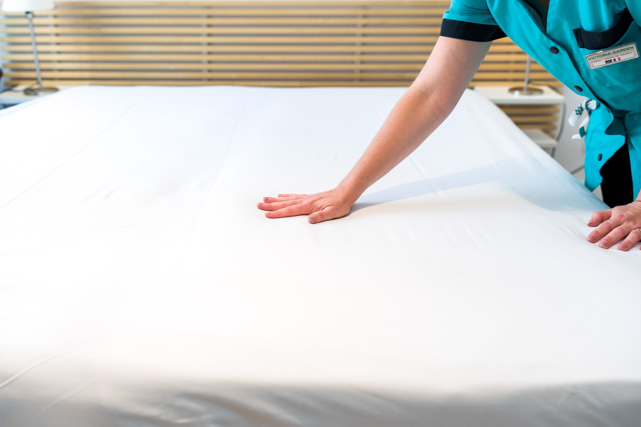 Une personne portant une chemise d'uniforme verte et un badge nominatif défroisse un drap blanc sur un lit soigneusement fait dans les Appartements De Vacances Bordeaux. La tête de lit est en bois avec des lattes horizontales et une partie d'une table de nuit avec une lampe de lecture intégrée est visible sur le côté gauche.