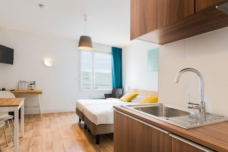 Ce studio moderne des Appartements De Vacances Bordeaux comprend une petite cuisine avec un évier en acier inoxydable et des meubles en bois. La chambre comprend un lit avec des draps blancs et des oreillers jaunes, un bureau et des chaises, une télévision murale et une fenêtre avec des rideaux bleus. Le parquet en bois clair met en valeur cet espace bien éclairé.