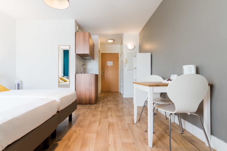 Un studio moderne et minimaliste des Appartements De Vacances Bordeaux dispose d'un lit avec des draps blancs, d'une petite cuisine avec des armoires en bois, d'une table à manger avec deux chaises blanches et d'un placard mural blanc. La pièce est dotée d'un parquet en bois clair, de murs blancs et gris et d'une abondante lumière naturelle provenant d'une fenêtre invisible.