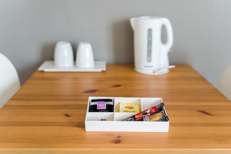 Dans l'espace cosy des Appartements De Vacances Bordeaux, une table en bois avec un plateau blanc contenant divers sachets de thé et de café trône. Derrière le plateau, une bouilloire électrique blanche et deux tasses blanches sont posées à l'envers sur un plateau blanc assorti, contre un mur gris uni.