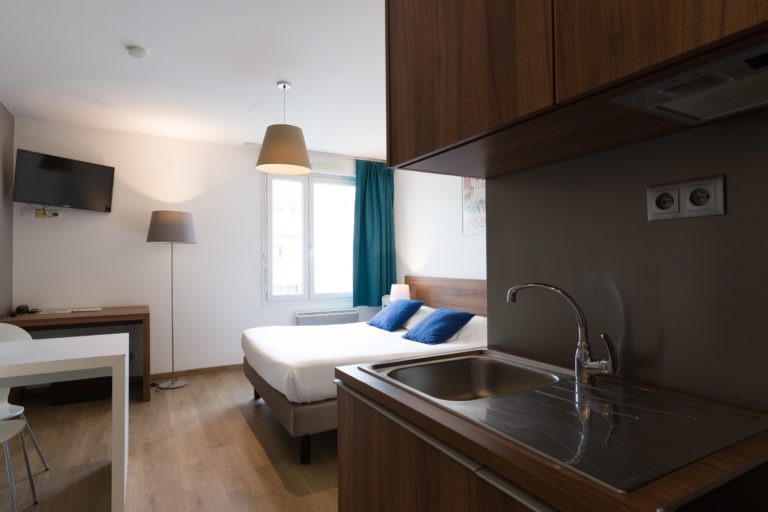 Studio moderne des Appartements De Vacances Bordeaux doté d'un lit bien fait avec des oreillers bleus, d'une télévision murale, d'un lampadaire et d'une fenêtre avec des rideaux bleu canard. Un bureau en bois est placé près de la fenêtre. Au premier plan, une petite kitchenette avec un évier, une cuisinière et des armoires en bois est visible.